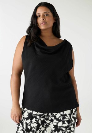 Donna che indossa una canotta nera senza maniche con scollo drappeggiato e pantaloni a fantasia floreale bianco e nero, in piedi davanti a uno sfondo semplice.