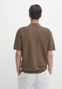 Polo shirt marron à manches courtes avec un motif tricoté texturé et un col doux. Le modèle porte un pantalon blanc, vu de dos.