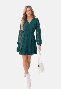 Abito verde in pizzo con maniche lunghe, design avvolgente e gonna a strati. Abbinato a stivaletti bianchi e una borsa bianca.