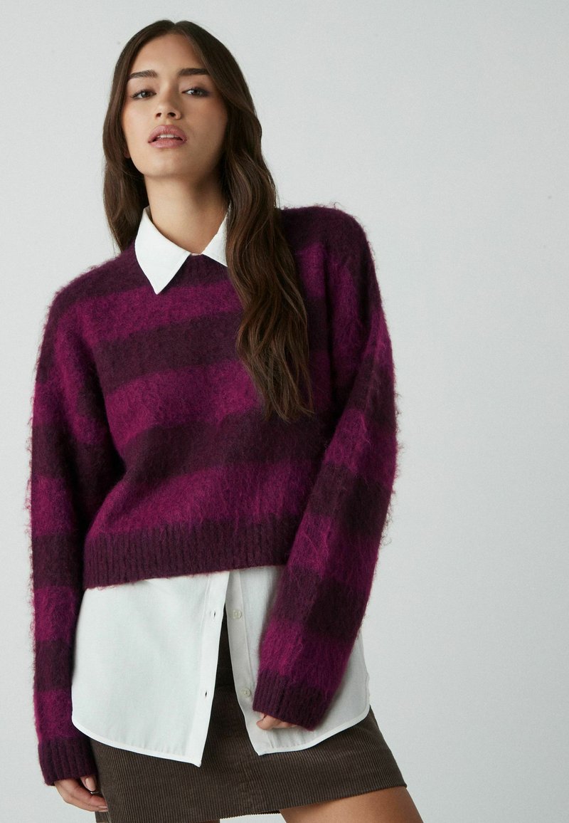 Sweater med lilla og mørke stribemønster, lavet af fluffy materiale, har en rund halsudskæring og løse ærmer over en hvid skjorte med krave.