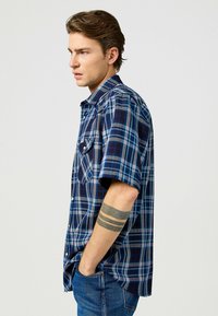 Camicia a maniche corte a quadri in blu scuro e blu con dettagli grigio chiaro e colletto con bottoni. Presenta due tasche sul petto e una vestibilità rilassata.