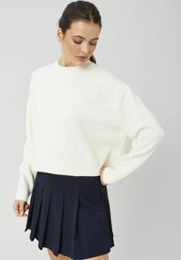 Femme aux cheveux foncés portant un pull en tricot blanc avec une broderie florale discrète et une jupe plissée bleu marine, debout devant un fond uni.