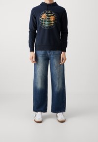 Sudadera con capucha azul marino con un estampado gráfico de palmeras, combinada con vaqueros de pierna ancha en azul y zapatillas blancas. Estilo casual y cómodo.