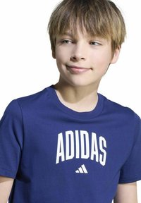 Jongen met lichtbruin haar, die een blauwe Adidas-t-shirt draagt, iets naar de zijkant kijkt met een neutrale uitdrukking.