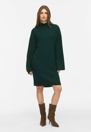 Woman standing wearing a dark green long-sleeve knitted dress with a high neck and brown suede mid-calf heeled boots.