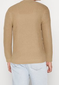 Suéter beige de punto con mangas largas y textura acanalada, con un escote redondo y dobladillo recto, llevado encima de unos vaqueros azul claro.