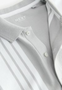 Polo rayé gris clair et blanc avec deux boutons blancs et un col côtelé, étiqueté "NEXT" fabriqué au Pakistan.