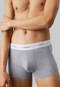 Sivi pamučni bokserice s bijelim pojasom na kojem piše "Calvin Klein". Tkanina ima mekanu teksturu i blizak kroj.