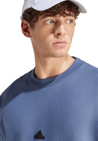 Felpa blu con scollatura a costine e logo Adidas nero sul petto. Il modello indossa un cappello bianco. Il tessuto appare morbido e liscio.