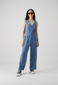 Denim kombinezon v svetlo modri barvi, z V-izrezom, gumbom spredaj, širokimi hlačami in teksturirano tkanino. Nosi se z črnimi sončnimi očali in supergami.