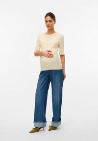 Top a costine crema con maniche corte, abbinato a jeans blu larghi e scarpe a punta verde oliva. In posa eretta con una mano sulla pancia.