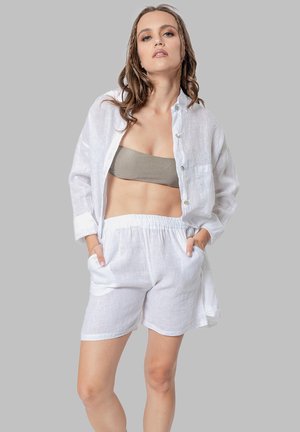Vrouw in lichtgewicht witte linnen shorts en open button-down blouse over een grijze bandeau, staand met handen in de zakken tegen een grijze achtergrond.