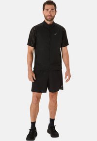Sort atletisk vest med lynlås fortil, uden ærmer, lavet af let materiale. Kombinationen inkluderer sorte shorts og sneakers.