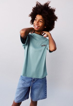 Garçon souriant aux cheveux bouclés portant un T-shirt turquoise avec un logo de bus et un short en jean, ajustant le col avec les mains derrière le cou.