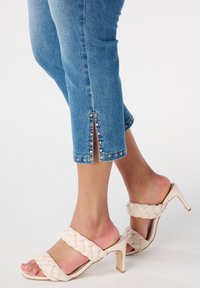 Pantalons capri en denim bleu clair avec des fentes sur les côtés et des clous argentés, associés à des sandales à talons en crème tissées avec un design de bride épaisse.