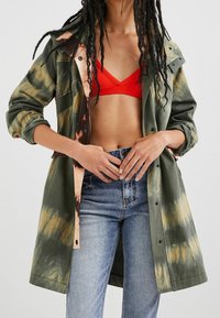 Giacca tie-dye verde oliva con un interno a contrasto marrone e pesca, chiusura con bottoni, vestibilità ampia e maniche lunghe, indossata sopra un bralette rosso.