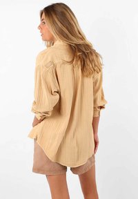 Chemise beige à boutons avec des rayures verticales, manches retroussées et ourlet arrondi, associée à un short marron clair. Détail subtil de l'étiquette de marque visible.