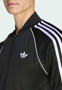 Άνδρας που φοράει μαύρο αθλητικό μπουφάν Adidas με λευκές ρίγες στους ώμους και έμβλημα στο στήθος, σε φωτεινό φόντο.