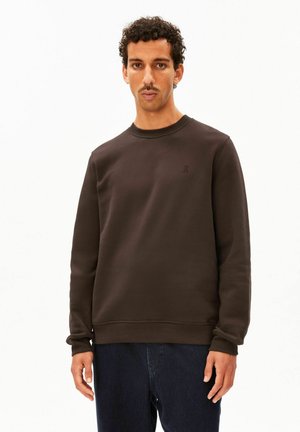 Mann mit kurzen lockigen Haaren und Schnurrbart, der ein dunkelbraunes Sweatshirt mit Rundhalsausschnitt und langen Ärmeln sowie dunkelblaue Jeans trägt und vor weißem Hintergrund steht.