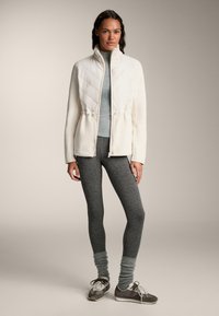 Jachetă albă matlasată cu talie ajustabilă prin cordon, purtată cu un top gri deschis și colanți gri închis. Pantofii sunt gri cu accente albe.