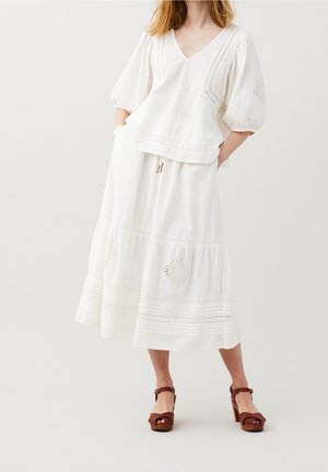 Kvinde iført hvid v-hals bluse med pufærmer og hvid broderet mellemlang nederdel, kombineret med brune platformsandaler.