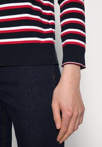 Navy-gestreifter Pullover mit roten und weißen horizontalen Streifen, gerippten Bündchen und einem weichen, strukturierten Stoff. Kombiniert mit dunklen Jeans.