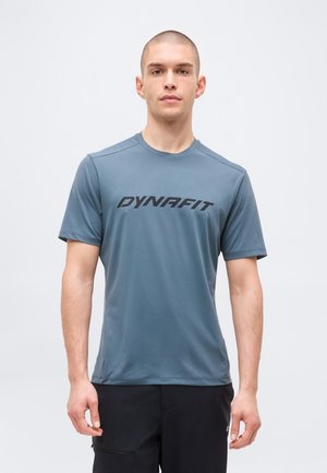 Νεαρός άνδρας με κοντά μαλλιά, φορώντας μπλε-γκρι μπλουζάκι με το λογότυπο Dynafit και μαύρο παντελόνι, στέκεται μπροστά σε απλό λευκό φόντο.