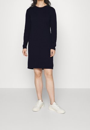 Person iført en sort kjole med lange ærmer og knælængde med hvide sneakers, der står mod en enkel lys baggrund.