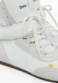 Bílé semišové vysoké tenisky s hladkým povrchem, tónovým prošíváním, polstrovaným pásem a žlutým logem. Tkaničky svázané vpředu.