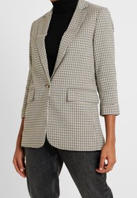 Prošedný béžový a černý blazer s jedním knoflíkem, klopami a dvěma předními kapsami; nošený přes černý žebrovaný rolák.