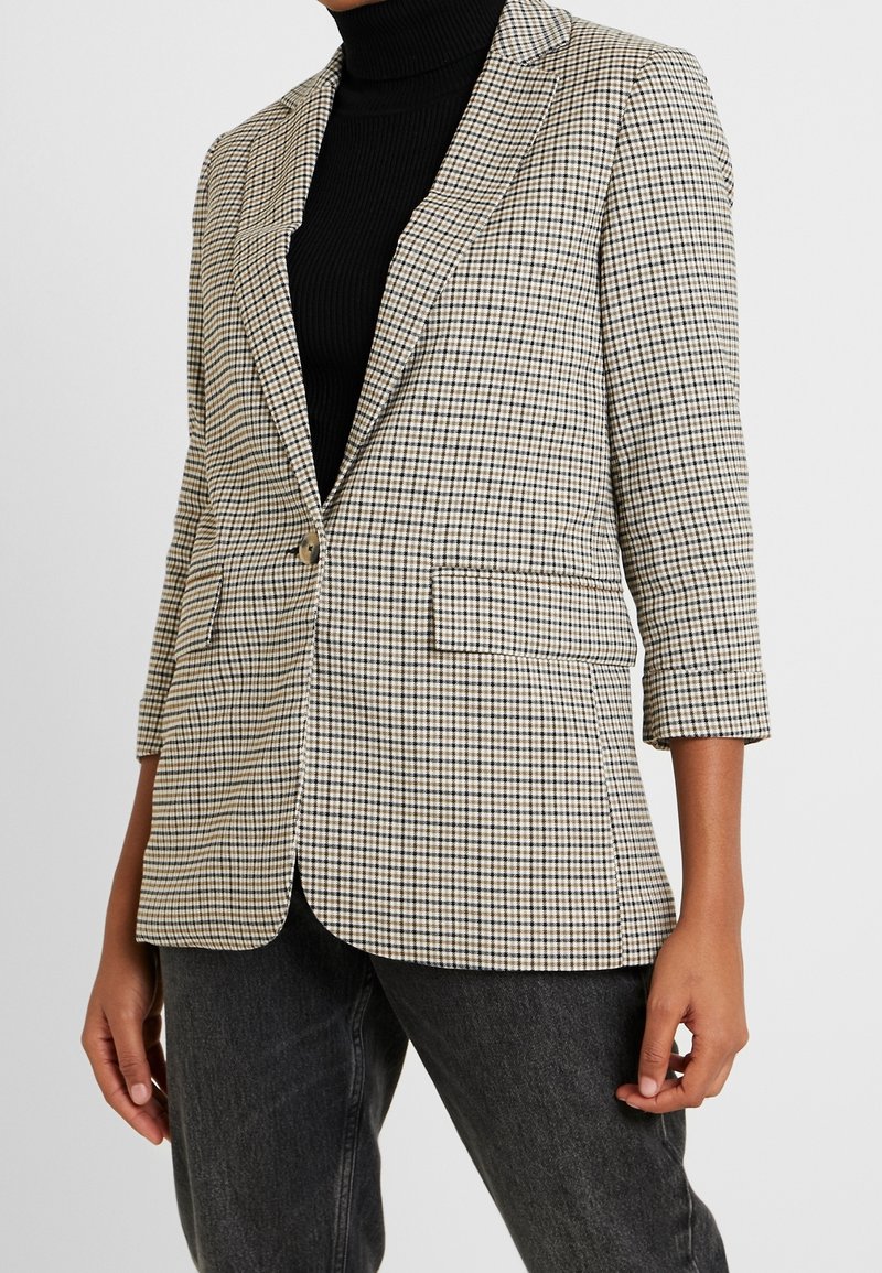 Prošedný béžový a černý blazer s jedním knoflíkem, klopami a dvěma předními kapsami; nošený přes černý žebrovaný rolák.