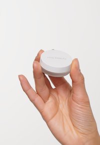 Vit cirkulär behållare som hålls i en hand, med en slät yta och präglad text "rms beauty" på locket.