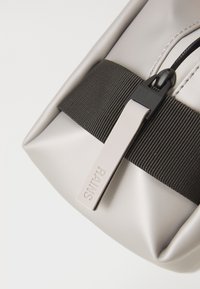 Lichtgrijze waterdichte tas met een glanzende afwerking, voorzien van een zwarte elastische band en een soepele rits met een logo label.
