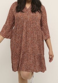Robe marron à motifs avec un décolleté en V, des manches trois-quarts et un design à niveaux. Le tissu semble léger et coule doucement.