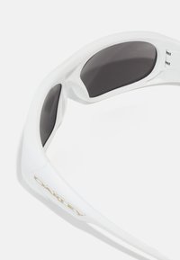 Białe okulary przeciwsłoneczne Oakley z szarymi soczewkami. Wygięta oprawka z acetatu ma gładką fakturę i złoty akcent logo z boku.