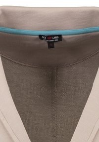 Beiger Kleidungsausschnitt mit einem sichtbaren schwarzen Etikett und blauen Akzenten im Kragen, das den Markennamen und die Größe klein zeigt.