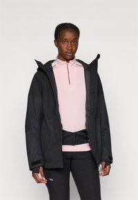 Casaco preto impermeável com capuz e painéis de tecido contrastantes, usado sobre uma camisa de mangas longas rosa clara, combinado com calças pretas.