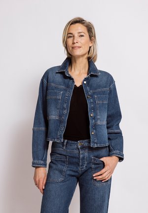 Frau mit kurzen blonden Haaren, die eine blaue Jeansjacke und Jeans trägt, steht mit einer Hand in der Tasche vor einem schlichten hellen Hintergrund.