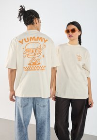 Beige oversized t-shirts with orange graphic print featuring a burger and text. One model wears faded blue jeans, the other black pants.