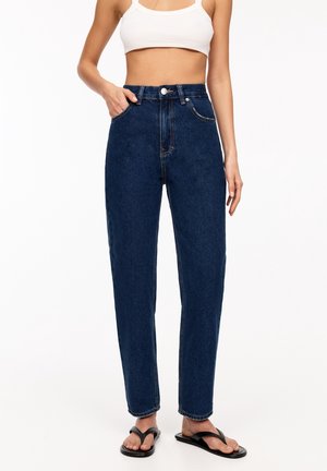 Femme portant un jean bleu foncé taille haute, un crop top blanc et des sandales noires, debout devant un fond blanc uni.