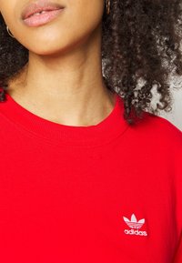 Czerwona bawełniana koszulka z krótkim rękawem, z ribbowanym dekoltem i małym białym logo Adidas wyhaftowanym po lewej stronie na klatce piersiowej.