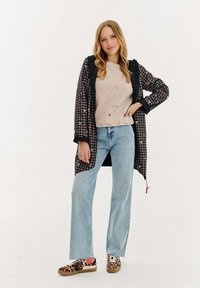 Sieviete, kas ģērbusies garā rūtainā mētelī ar sirdīm, smilšu krāsā drukātu džemperī, gaiši zilos džinsos un leoparda raksta kedās, stāv relaksēta.