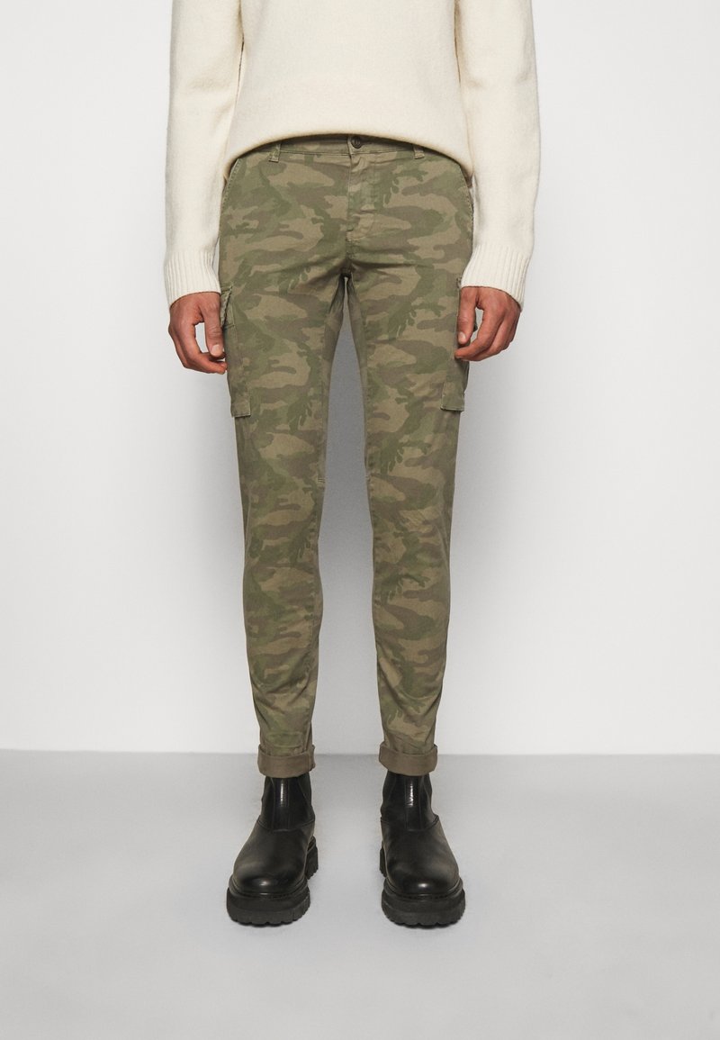 Kamouflage cargobyxor i gröna nyanser med lapplåtar, avsmalnande ben och uppvikt fåll, kombinerat med svarta boots som slutar vid anklarna.