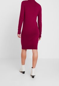 Åtsittande bordeauxfärgad klänning med hög halsringning och långa ärmar, tillverkad av ett mjukt stickat tyg, bärs med vita ankelstövlar.