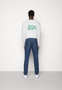 Sweatshirt cinza com texto verde "Quattro Stagioni" nas costas, combinado com jeans de denim azuis e ténis brancos, visto de costas.