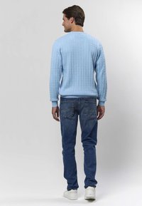 Lyseblå kabelstrikket genser med ribbestrikkede mansjetter, kombinert med mørke denimjeans og hvite sneakers, sett bakfra.