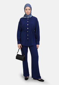 Navyblauwe twee-delige set bestaande uit een knoopsluiting top met een elastische taille en lange mouwen, gecombineerd met flared broeken. Accentuatie met een gekleurde hoofddoek.