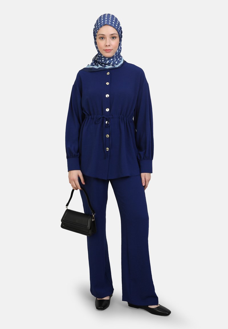 Navyblauwe twee-delige set bestaande uit een knoopsluiting top met een elastische taille en lange mouwen, gecombineerd met flared broeken. Accentuatie met een gekleurde hoofddoek.