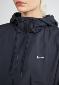 Musta nailonitakki hupulla, edessä vetoketju ja kiristysnyörit. Oikeassa rintapalassa pieni valkoinen Nike-logo. Sujuva pinta.
