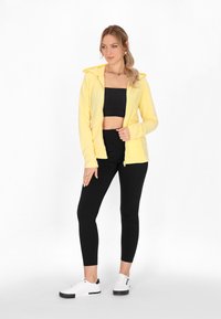Felpa gialla con zip e cappuccio, indossata sopra un crop top nero e leggings neri. Sneakers bianche con dettagli neri completano l'outfit.